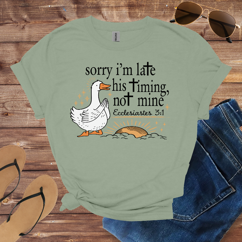 Green t-shirt with a goose graphic and text on a wooden surface with sunglasses and jeans.