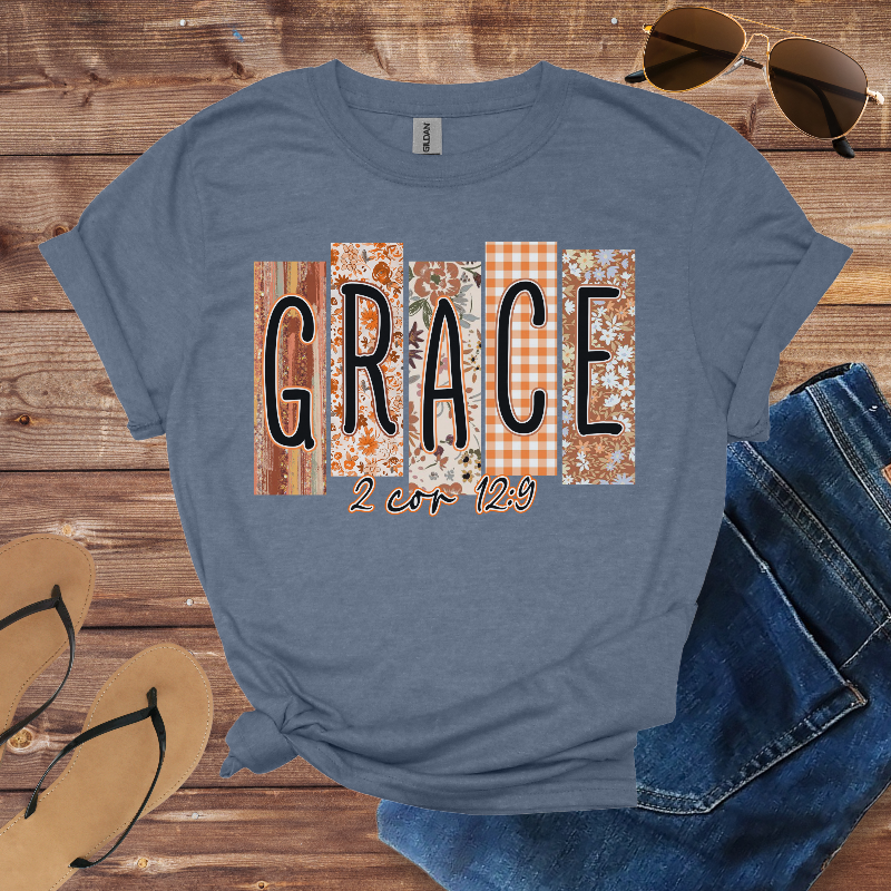 Blue t-shirt with 'GRACE 2 Cor 12:9' design on a wooden surface with sunglasses, jeans, and sandals.