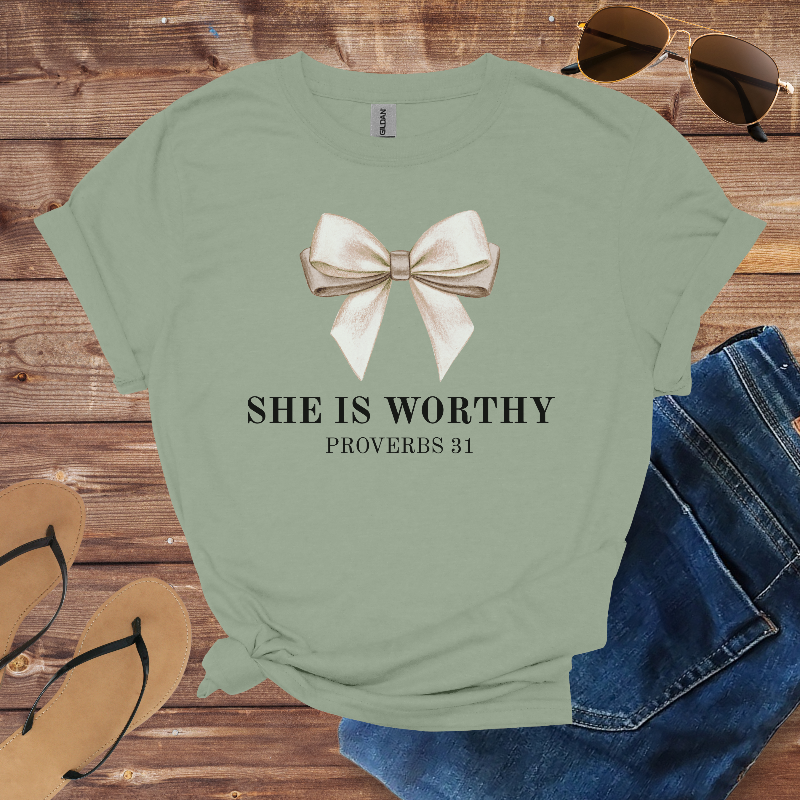 Green t-shirt with a bow graphic and 'SHE IS WORTHY Proverbs 31' text on a wooden surface with sunglasses, jeans, and sandals.