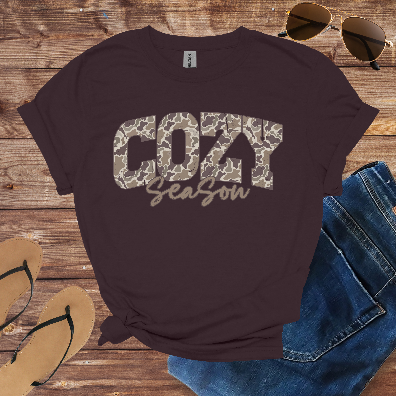 Brown t-shirt with 'COZY Season' text on a wooden surface with sunglasses, jeans, and flip-flops.