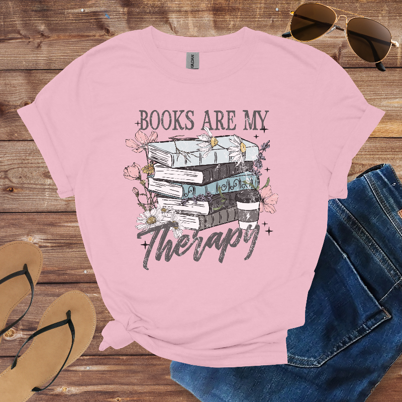 Pink t-shirt with 'Books are my Therapy' design on a wooden surface with sunglasses, jeans, and sandals.