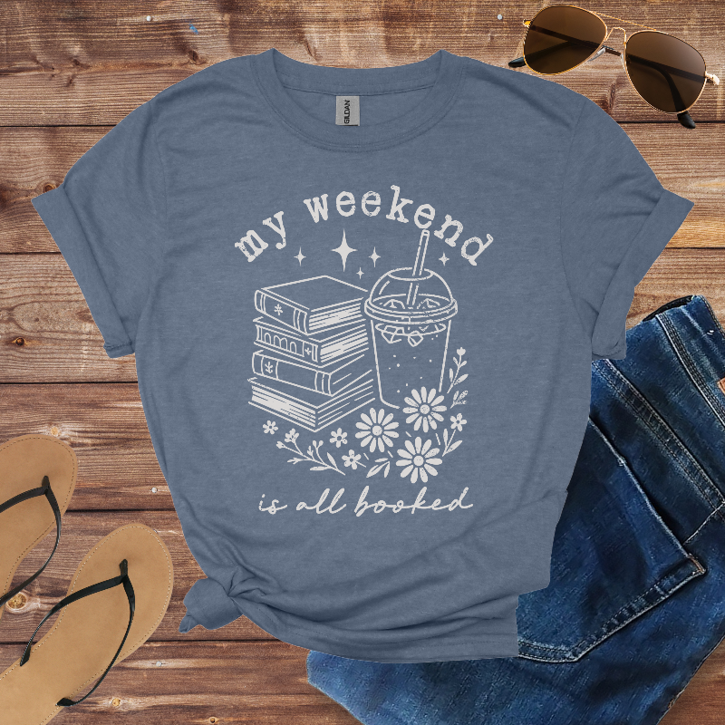 Blue t-shirt with 'my weekend is all booked' design on a wooden surface with sunglasses, jeans, and sandals.