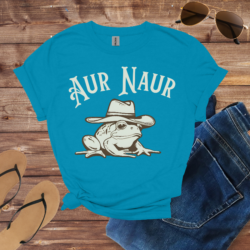 Blue t-shirt with 'Aur Naur' and frog graphic on a wooden surface with sunglasses and jeans.