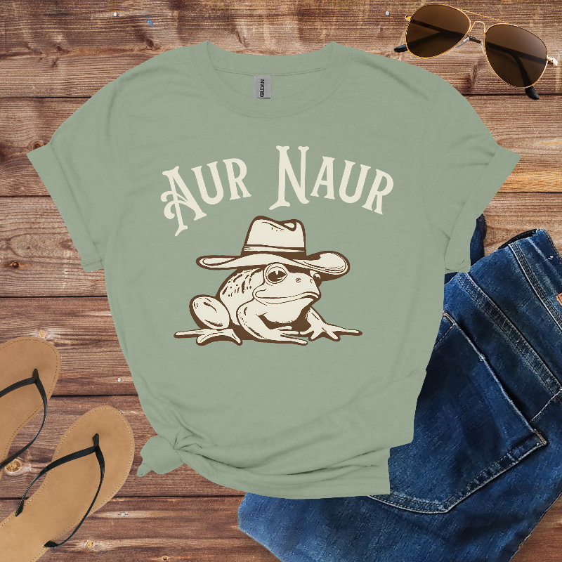 Green t-shirt with a frog wearing a hat graphic and 'Aur Naur' text, placed on a wooden surface with sunglasses and jeans.