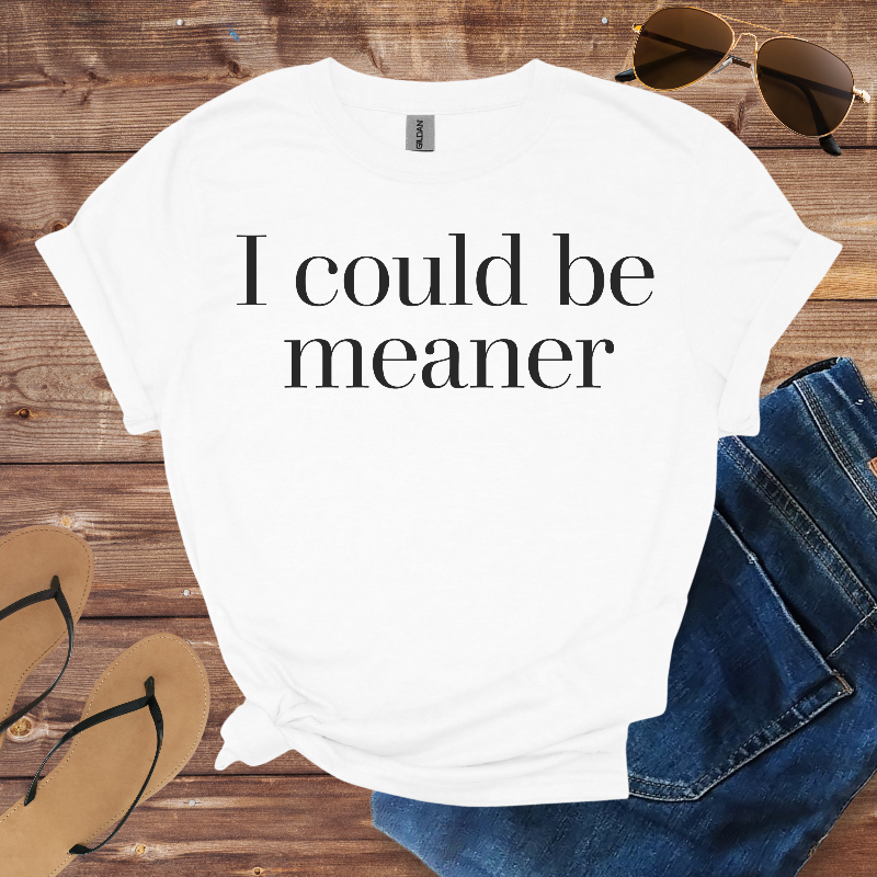 White t-shirt with 'I could be meaner' text on a wooden surface with sunglasses, jeans, and sandals.