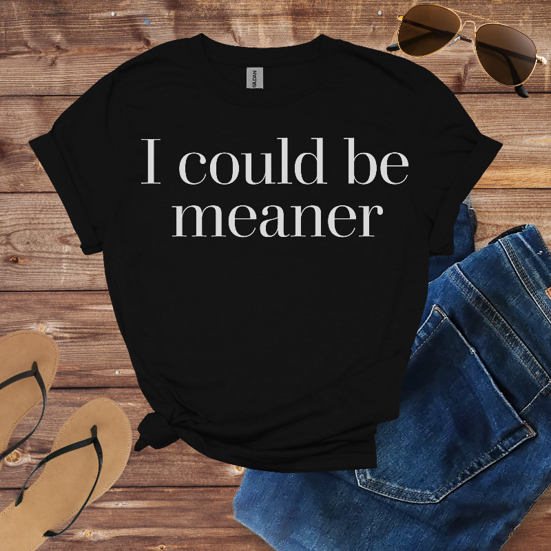 Black t-shirt with 'I could be meaner' text on a wooden surface with sunglasses, jeans, and sandals.