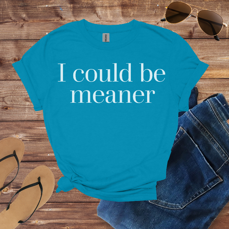 Blue t-shirt with 'I could be meaner' text on a wooden surface with sunglasses, jeans, and sandals.