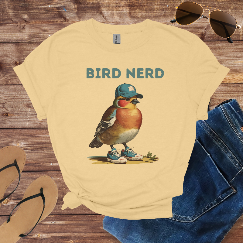 Beige t-shirt with a bird graphic and 'BIRD NERD' text on a wooden surface with sunglasses, jeans, and flip-flops.