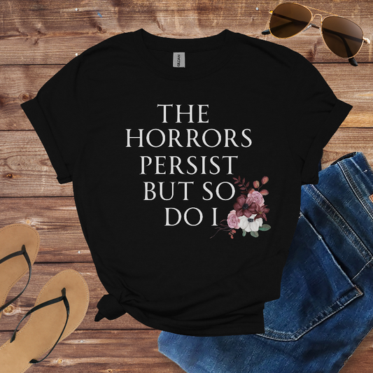 Black t-shirt with 'The Horrors Persist But So Do I' text and floral design on a wooden surface with sunglasses, jeans, and sandals.