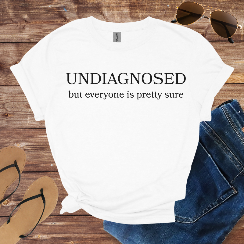 White t-shirt with 'UNDIAGNOSED but everyone is pretty sure' text on a wooden surface with jeans, sunglasses, and flip-flops.