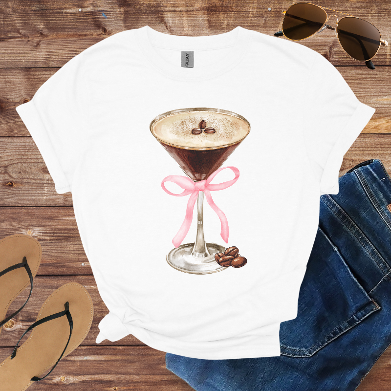 White t-shirt with a cocktail graphic on a wooden surface with sunglasses and jeans.