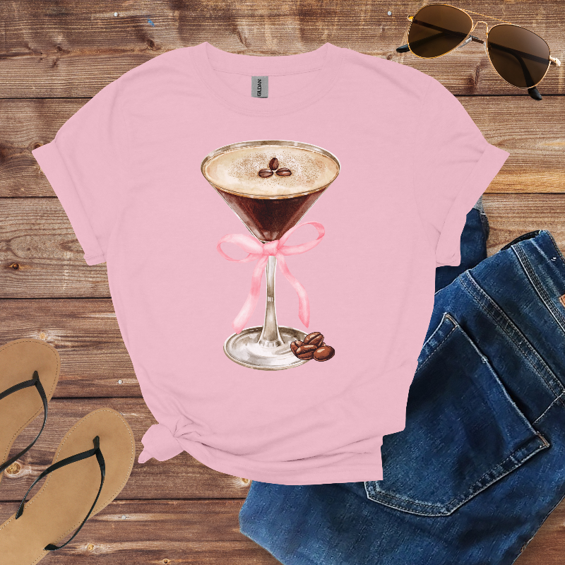 Pink t-shirt with a coffee-themed design on a wooden surface with sunglasses and jeans.