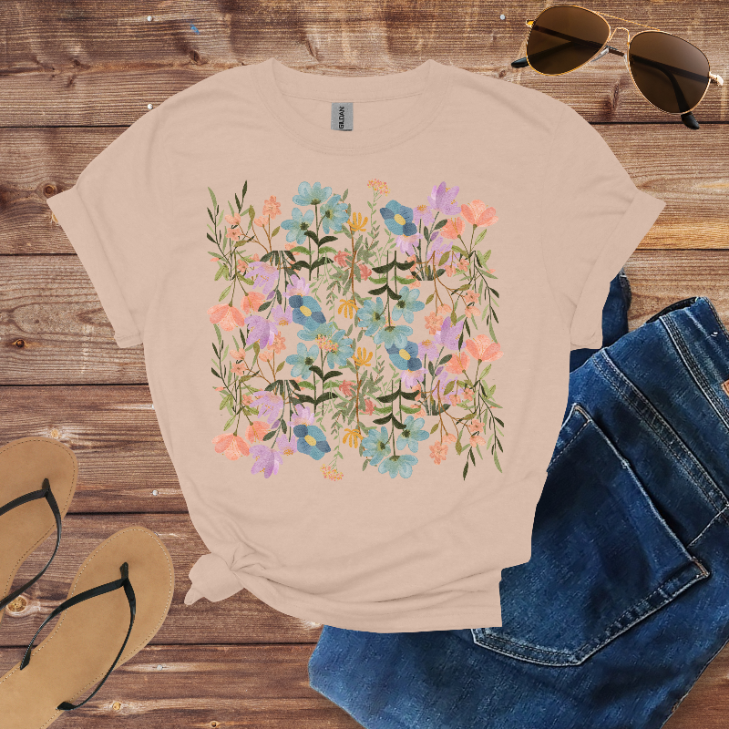 Beige t-shirt with floral design on a wooden surface with sunglasses, jeans, and sandals.