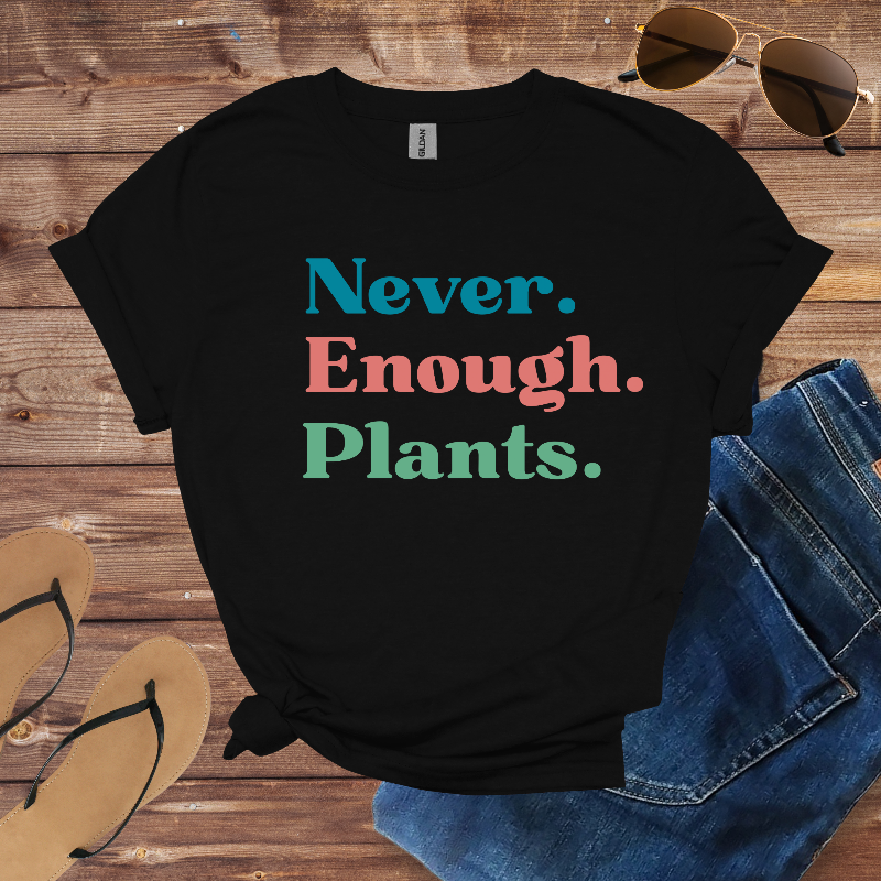 Black t-shirt with 'Never. Enough. Plants.' text on a wooden surface with sunglasses, jeans, and sandals.