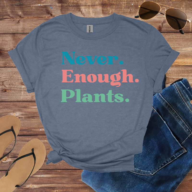Blue t-shirt with 'Never. Enough. Plants.' text on a wooden surface with sunglasses, jeans, and sandals.