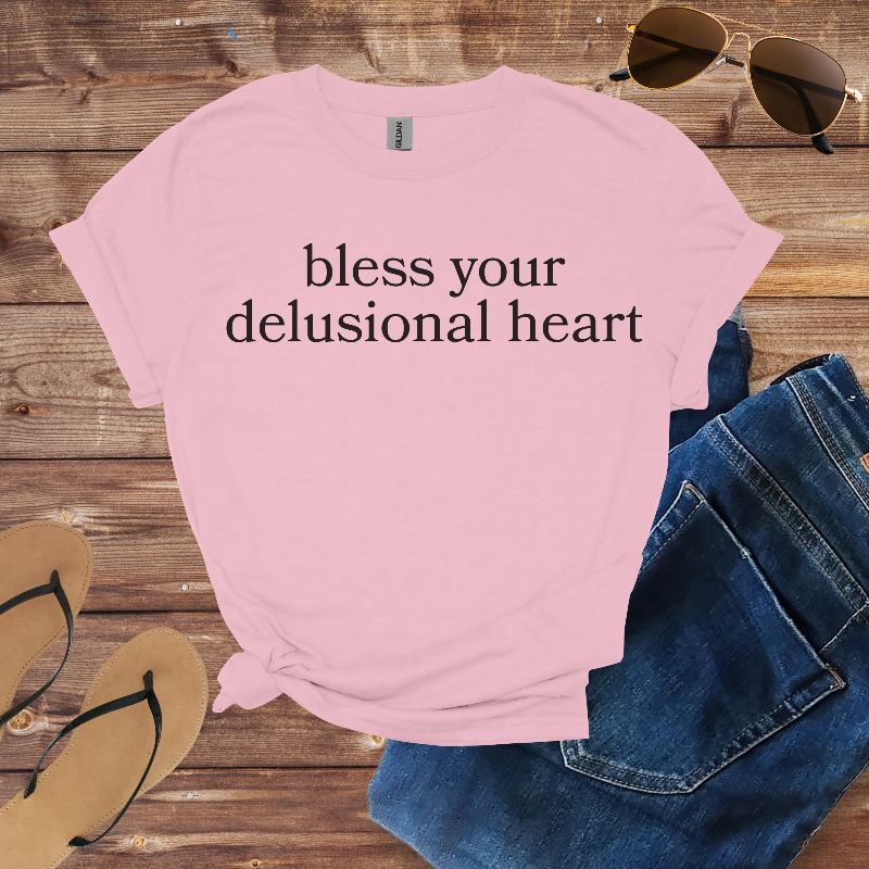 Pink t-shirt with text 'bless your delusional heart' on a wooden surface with sunglasses and jeans.