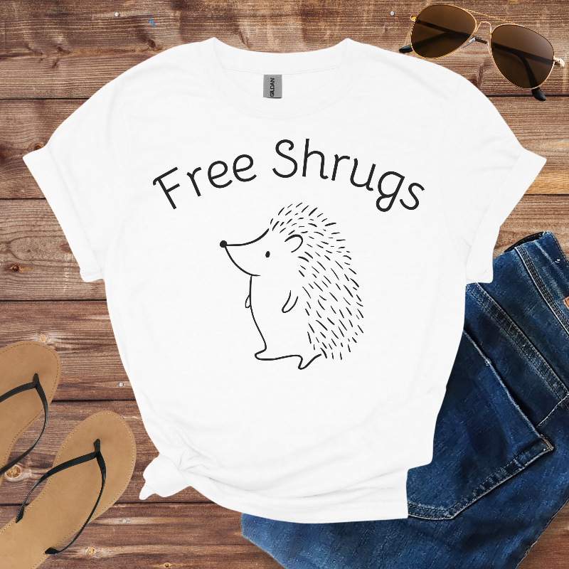 White t-shirt with 'Free Shrugs' text and hedgehog graphic on a wooden surface with sunglasses, jeans, and sandals.