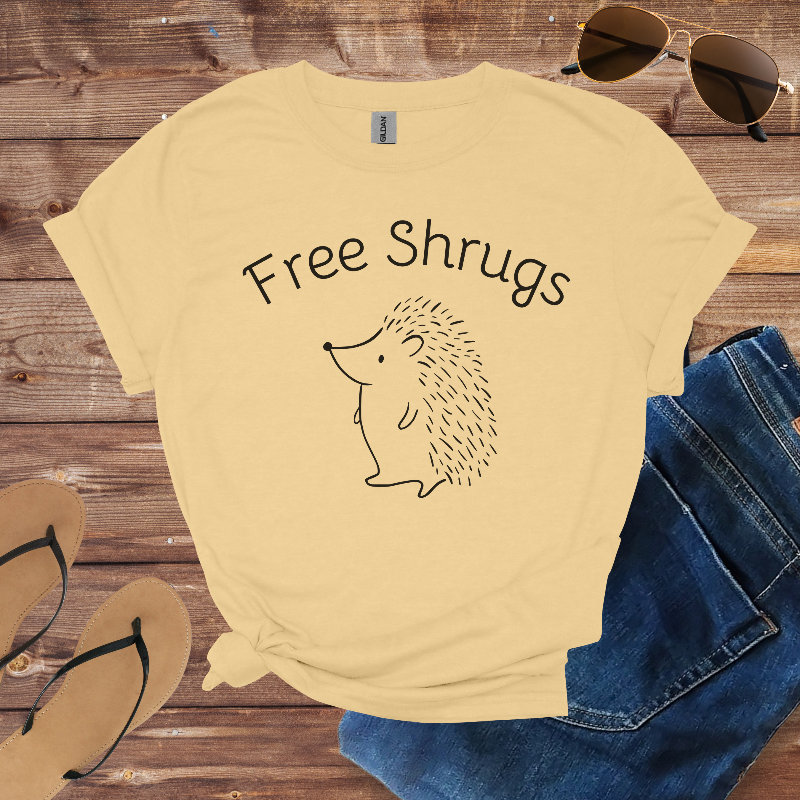 Beige t-shirt with 'Free Shrugs' text and hedgehog graphic on a wooden surface with sunglasses and jeans.