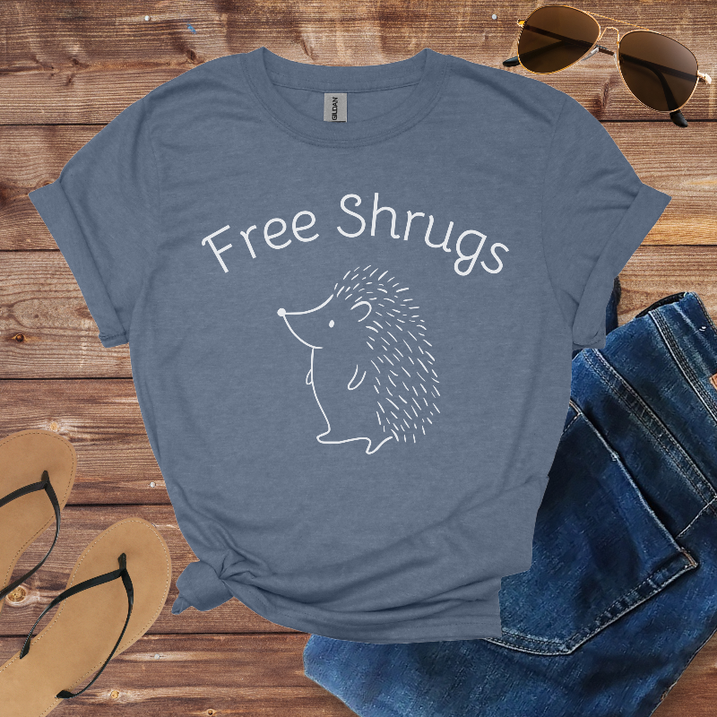 Blue t-shirt with 'Free Shrugs' text and hedgehog graphic on a wooden surface with sunglasses, jeans, and sandals.