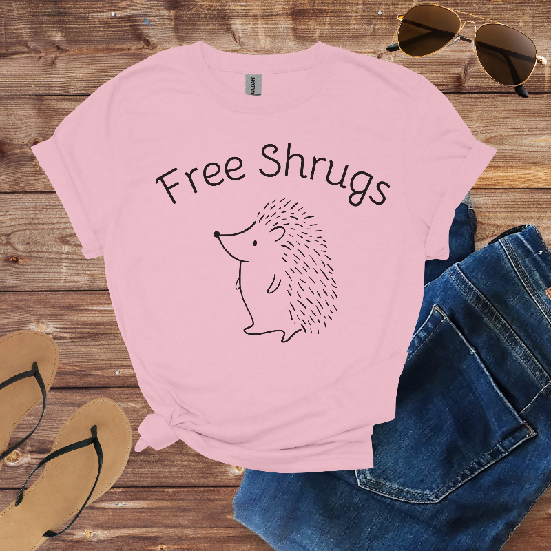 Pink t-shirt with 'Free Shrugs' text and hedgehog graphic on a wooden surface with sunglasses and sandals.