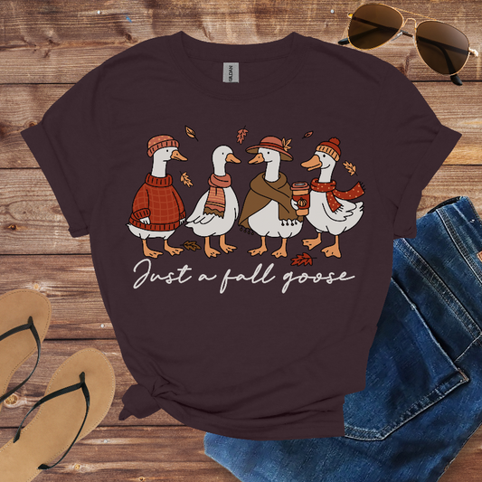 Dark brown t-shirt with cartoon geese design and text 'Just a fall goose' on a wooden surface with sunglasses, jeans, and sandals.