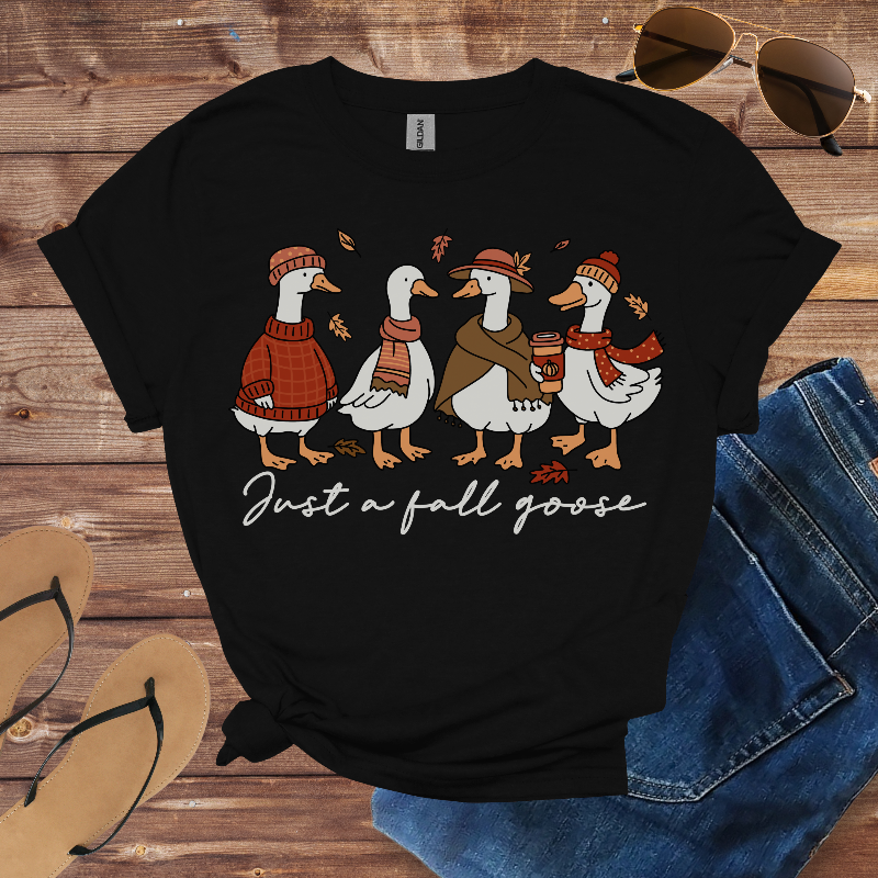 Black t-shirt with a graphic of ducks wearing fall-themed accessories and text 'Just a fall goose' on a wooden surface with jeans and sunglasses.