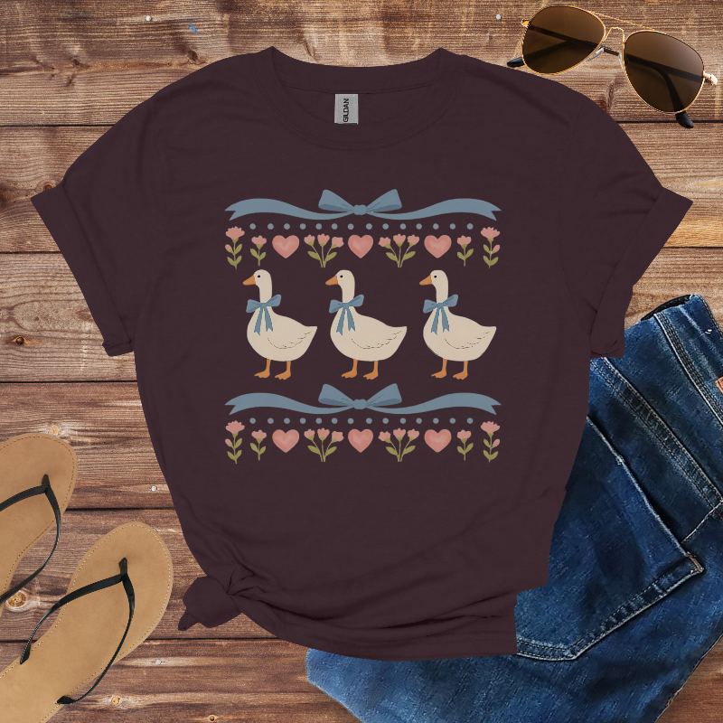 Dark purple t-shirt with duck design on a wooden surface with jeans, sandals, and sunglasses.