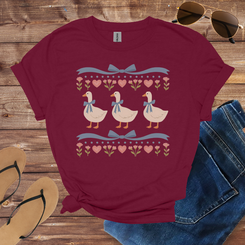 Maroon t-shirt with duck design on a wooden surface with sunglasses, jeans, and sandals.