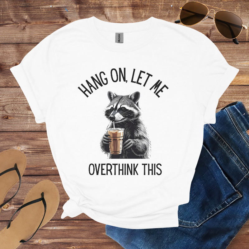 White t-shirt with raccoon graphic and text on a wooden surface with sunglasses and jeans.
