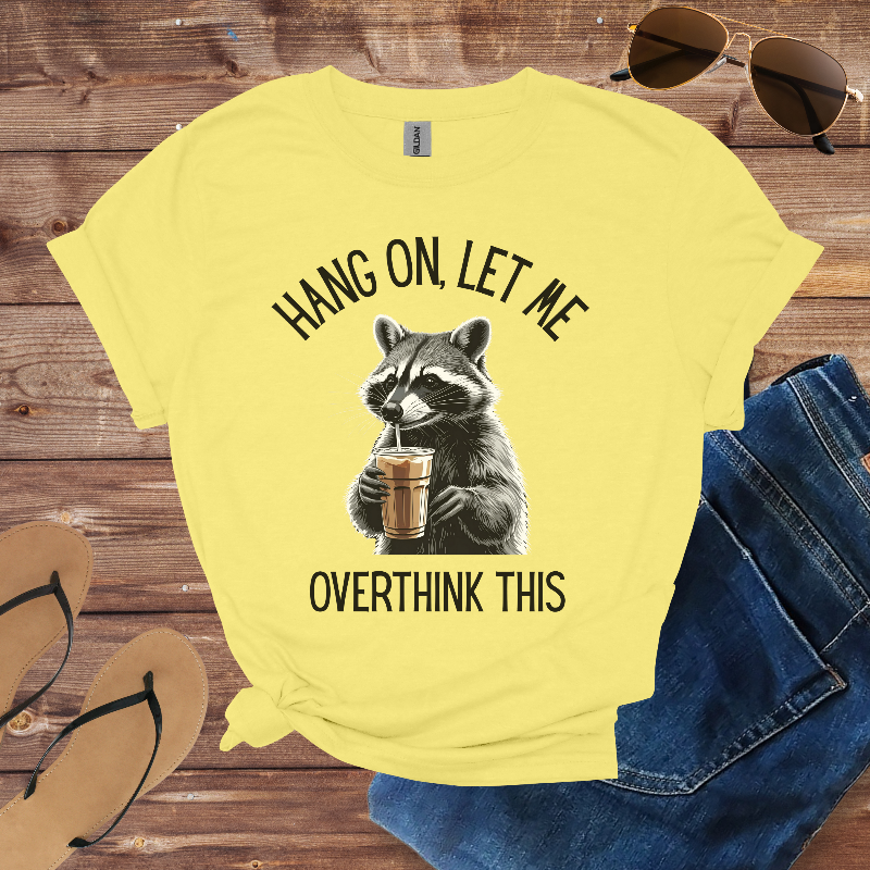 Yellow t-shirt with raccoon graphic and text on a wooden surface with sunglasses and jeans.
