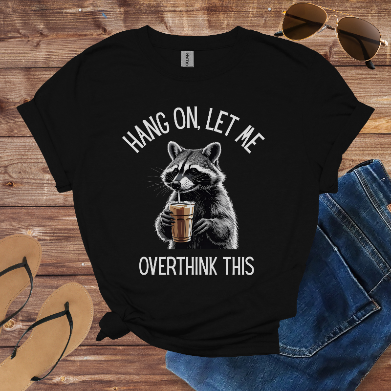 Black t-shirt with raccoon graphic and text on a wooden surface with sunglasses and jeans.