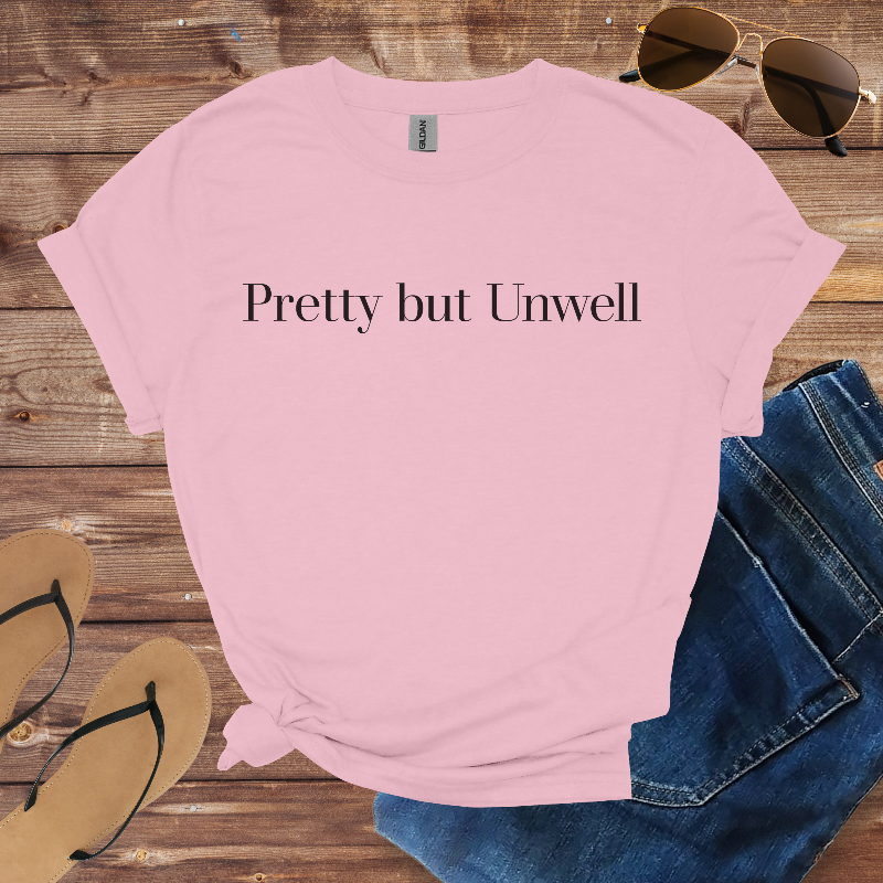 Pink t-shirt with 'Pretty but Unwell' text on a wooden surface with sunglasses, jeans, and flip-flops.