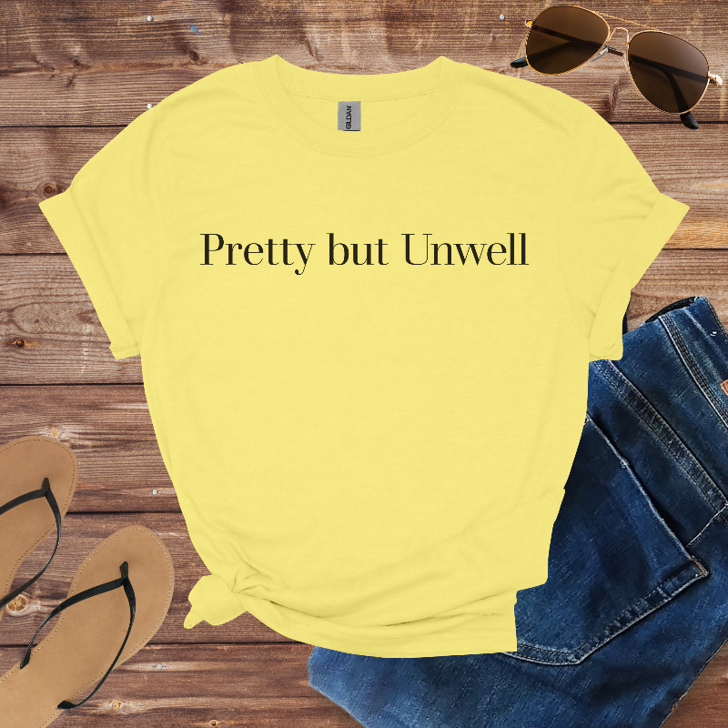 Yellow t-shirt with 'Pretty but Unwell' text on a wooden surface with sunglasses, jeans, and flip-flops.