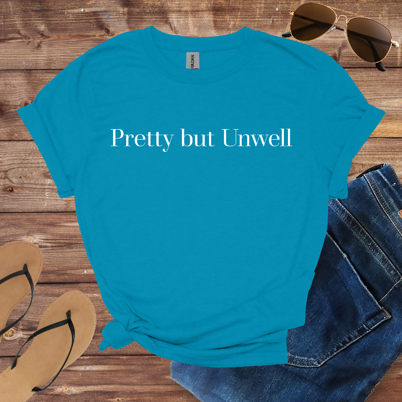 Blue t-shirt with 'Pretty but Unwell' text on a wooden surface with sunglasses, jeans, and sandals.