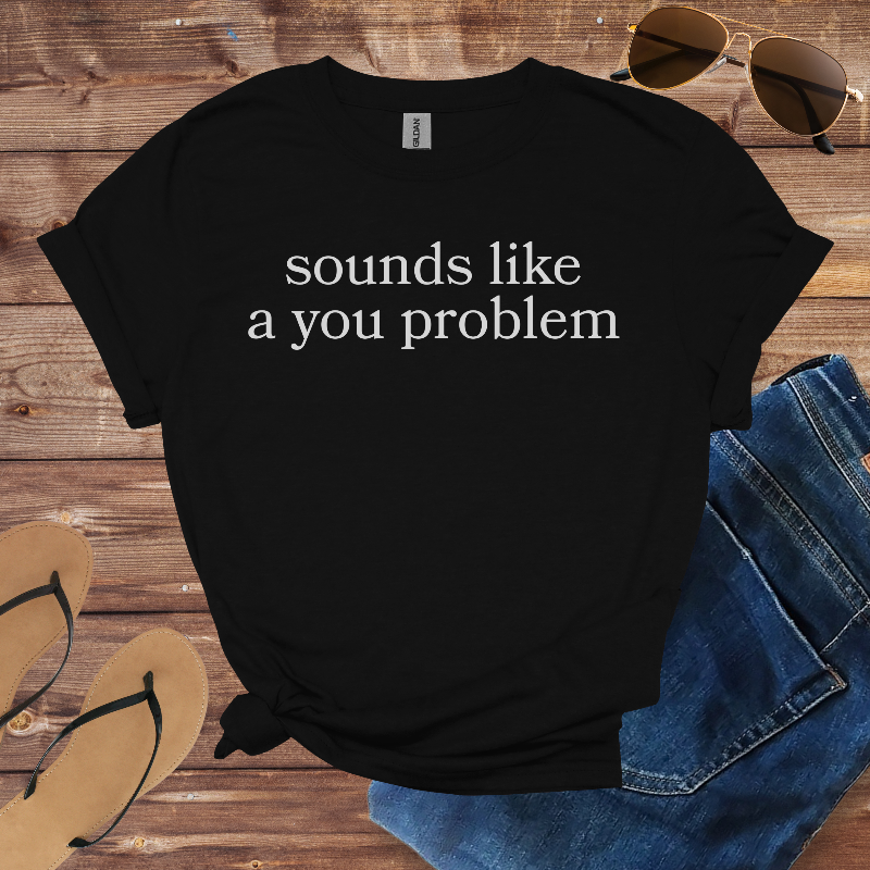 Black t-shirt with 'sounds like a you problem' text on a wooden surface with sunglasses, jeans, and sandals.
