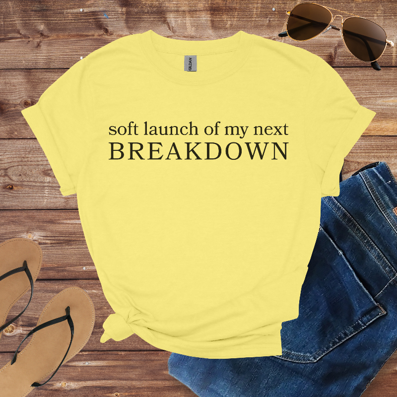 Yellow t-shirt with text 'soft launch of my next BREAKDOWN' on a wooden surface with jeans, sunglasses, and flip-flops.