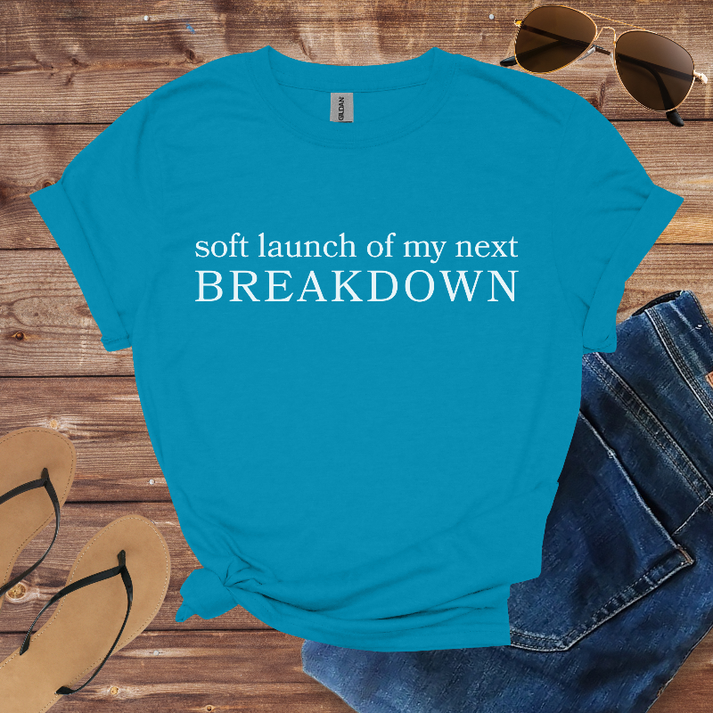 Blue t-shirt with humorous text on a wooden surface with sunglasses, jeans, and flip-flops.