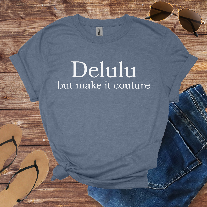Blue t-shirt with 'Delulu but make it couture' text on a wooden surface with sunglasses, jeans, and sandals.