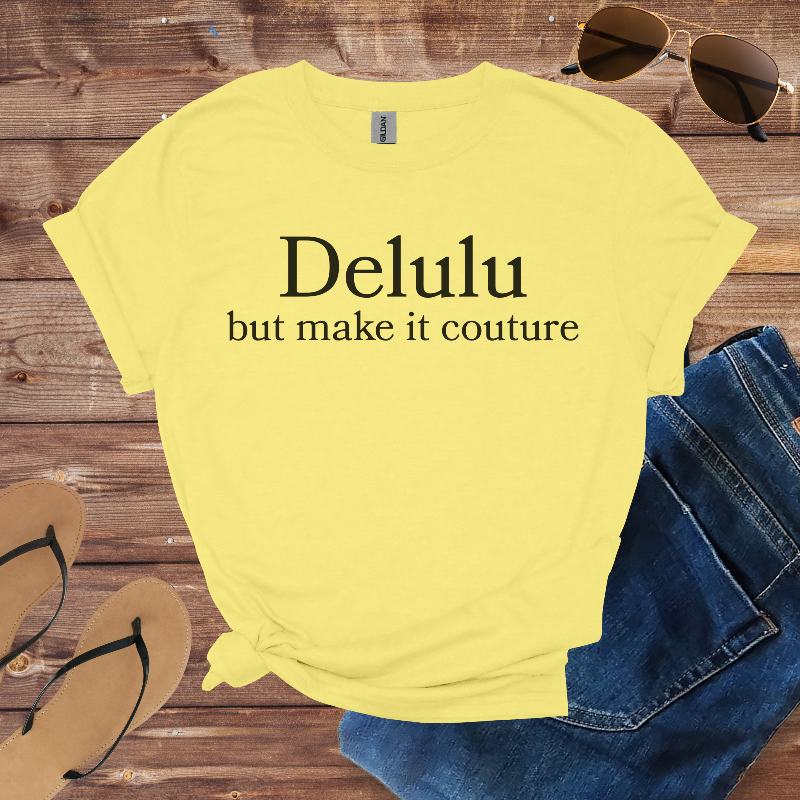 Yellow t-shirt with 'Delulu but make it couture' text on a wooden surface with sunglasses, jeans, and sandals.