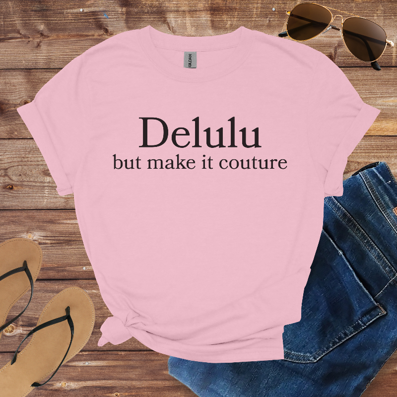 Pink t-shirt with 'Delulu but make it couture' text on a wooden surface with sunglasses and jeans.