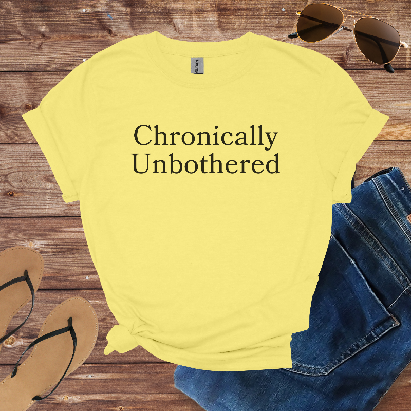 Yellow t-shirt with 'Chronically Unbothered' text on a wooden surface with sunglasses, jeans, and flip-flops.