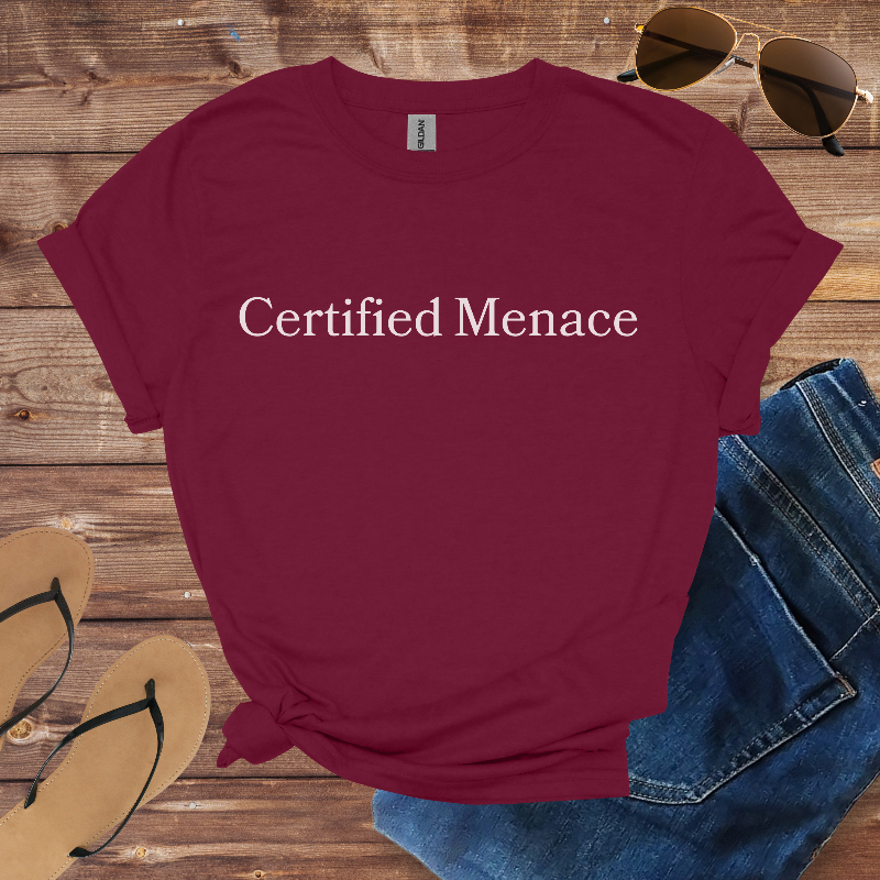 Maroon t-shirt with 'Certified Menace' text on a wooden surface with sunglasses, jeans, and sandals.