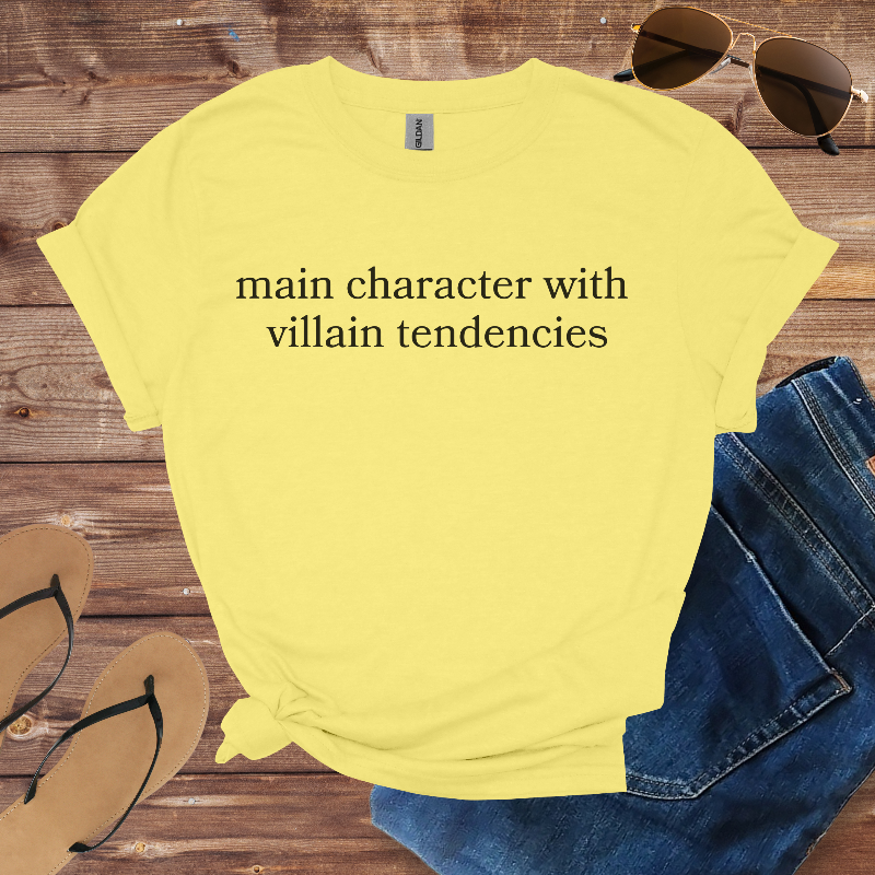 Yellow t-shirt with text 'main character with villain tendencies' on a wooden surface with sunglasses, jeans, and sandals.