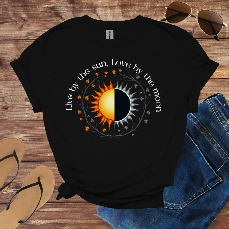 Black t-shirt with sun and moon design and text 'Live by the sun, Love by the moon' on a wooden surface with sunglasses and flip-flops.