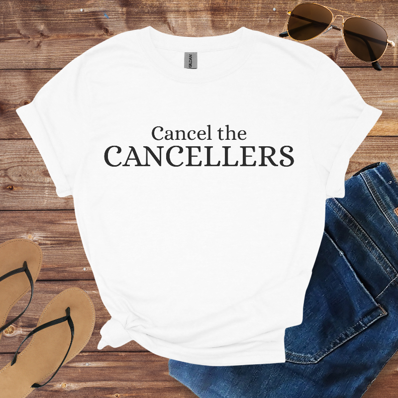 White t-shirt with 'Cancel the CANCELLERS' text on a wooden surface with sunglasses, jeans, and sandals.