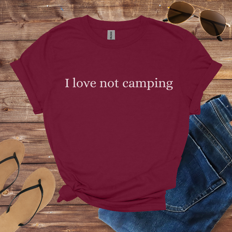 Maroon t-shirt with 'I love not camping' text on a wooden surface with sunglasses, flip-flops, and jeans.