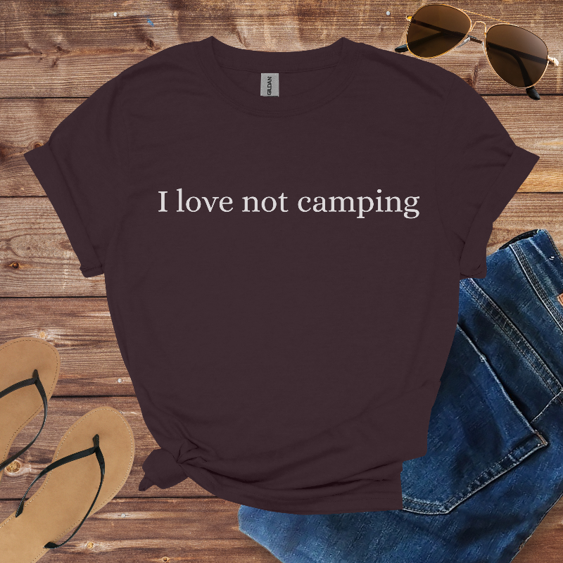 Brown t-shirt with 'I love not camping' text on a wooden surface with sunglasses, flip-flops, and jeans.