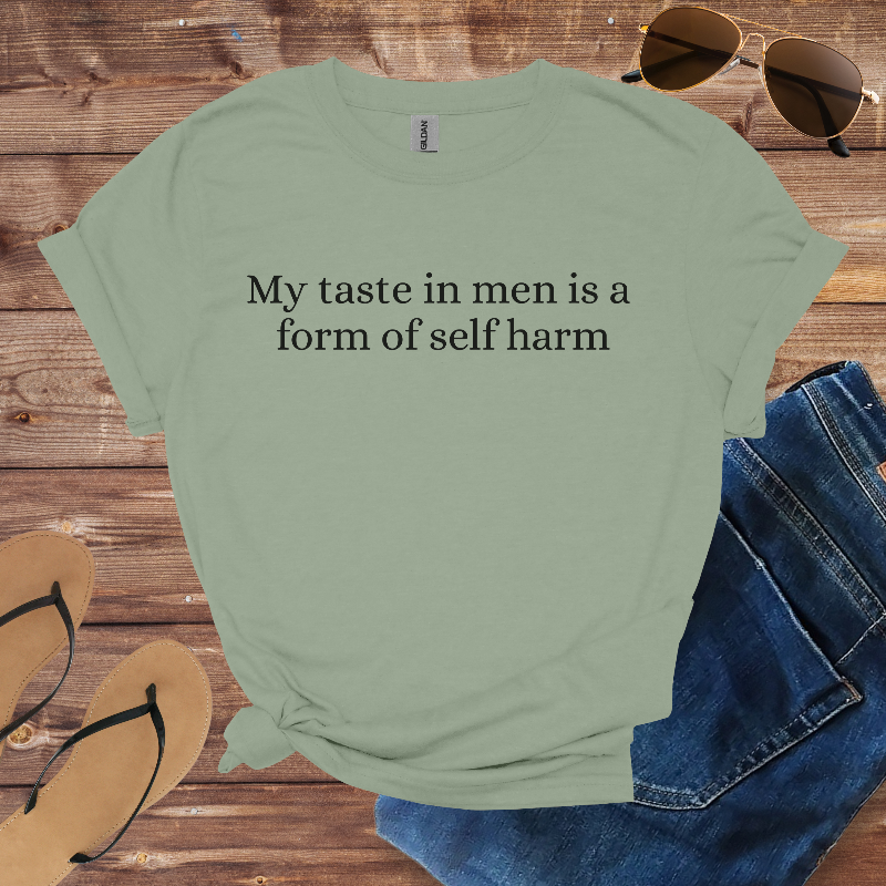 Green t-shirt with text 'My taste in men is a form of self harm' on a wooden surface with jeans, sandals, and sunglasses.