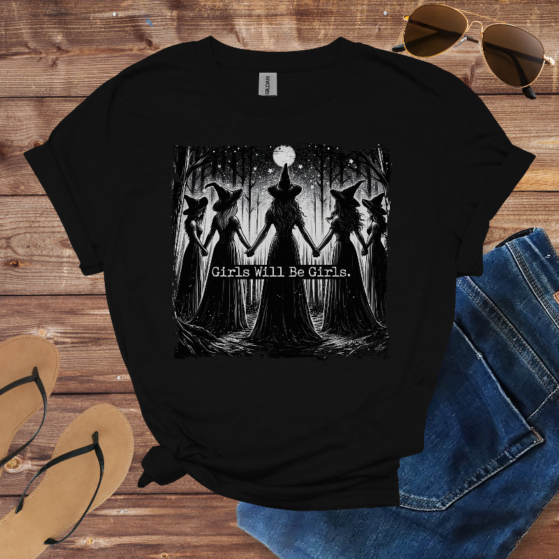Black t-shirt with witch graphic and 'Girls Will Be Girls' text on a wooden surface with sunglasses, flip-flops, and jeans.
