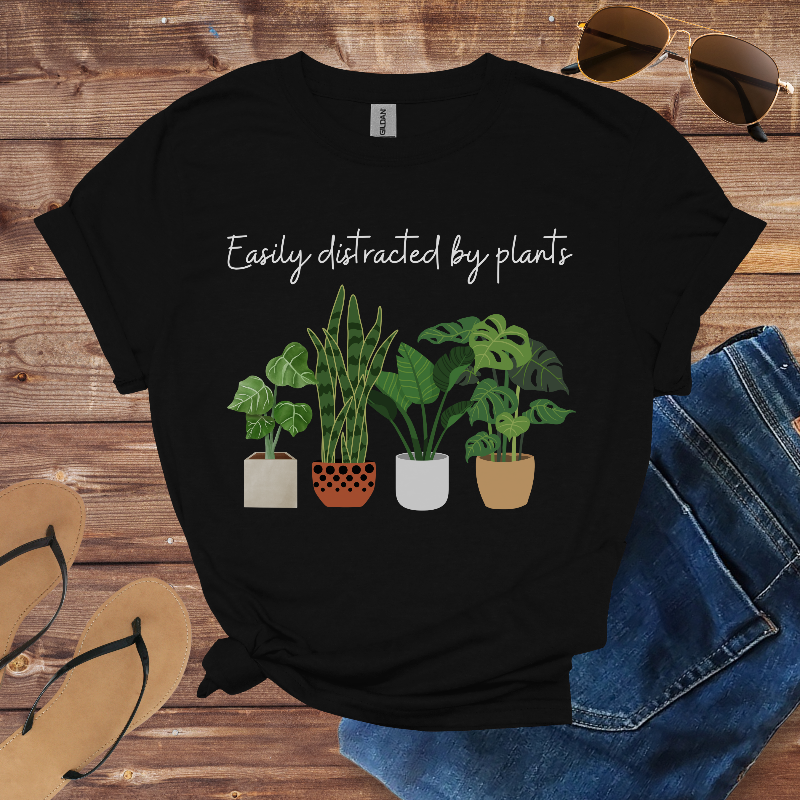 Black t-shirt with plant design and text 'Easily distracted by plants' on a wooden surface with sunglasses, jeans, and sandals.